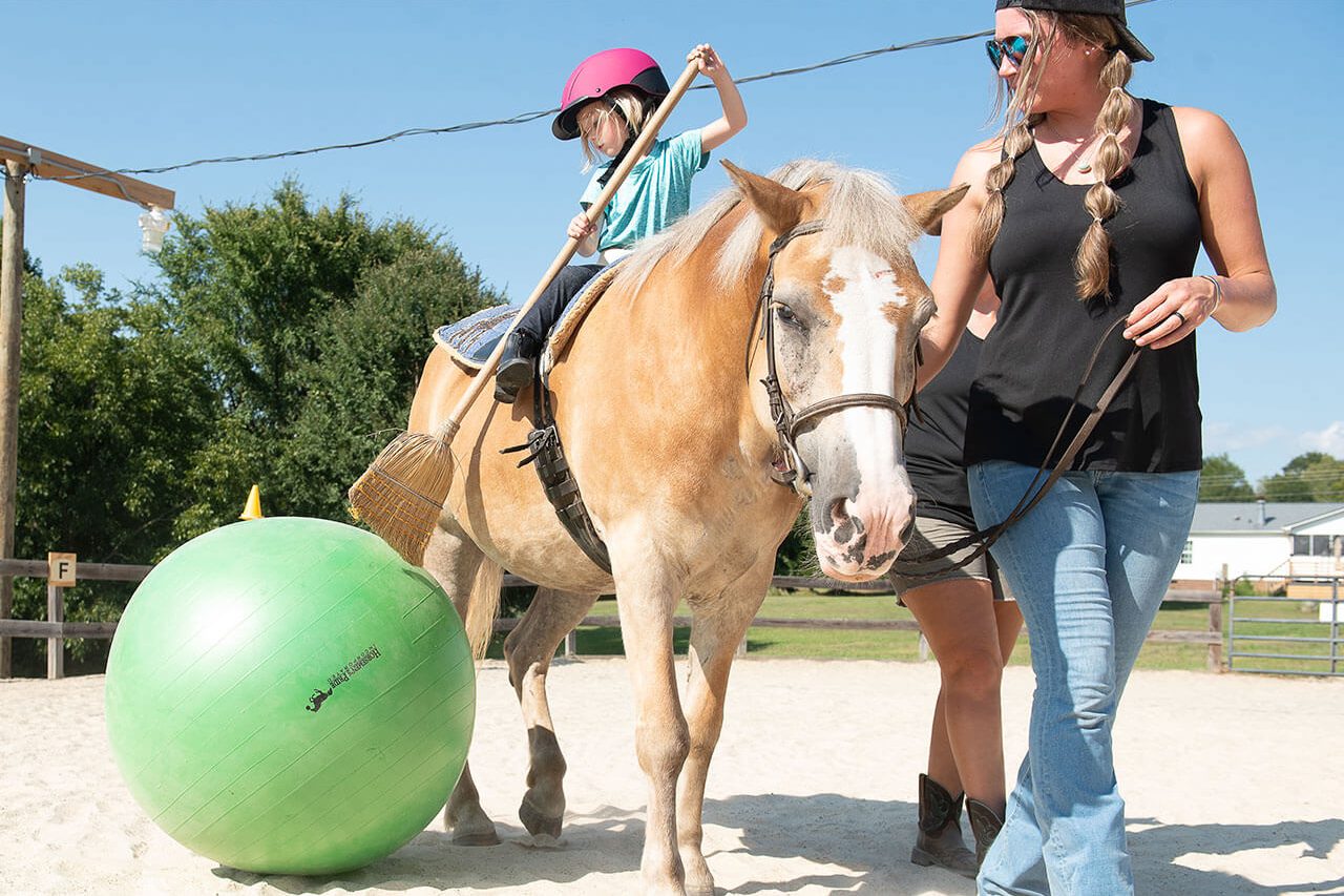 A young girl pushes an inflatable ball with a broom while on horseback during a hippotherapy session