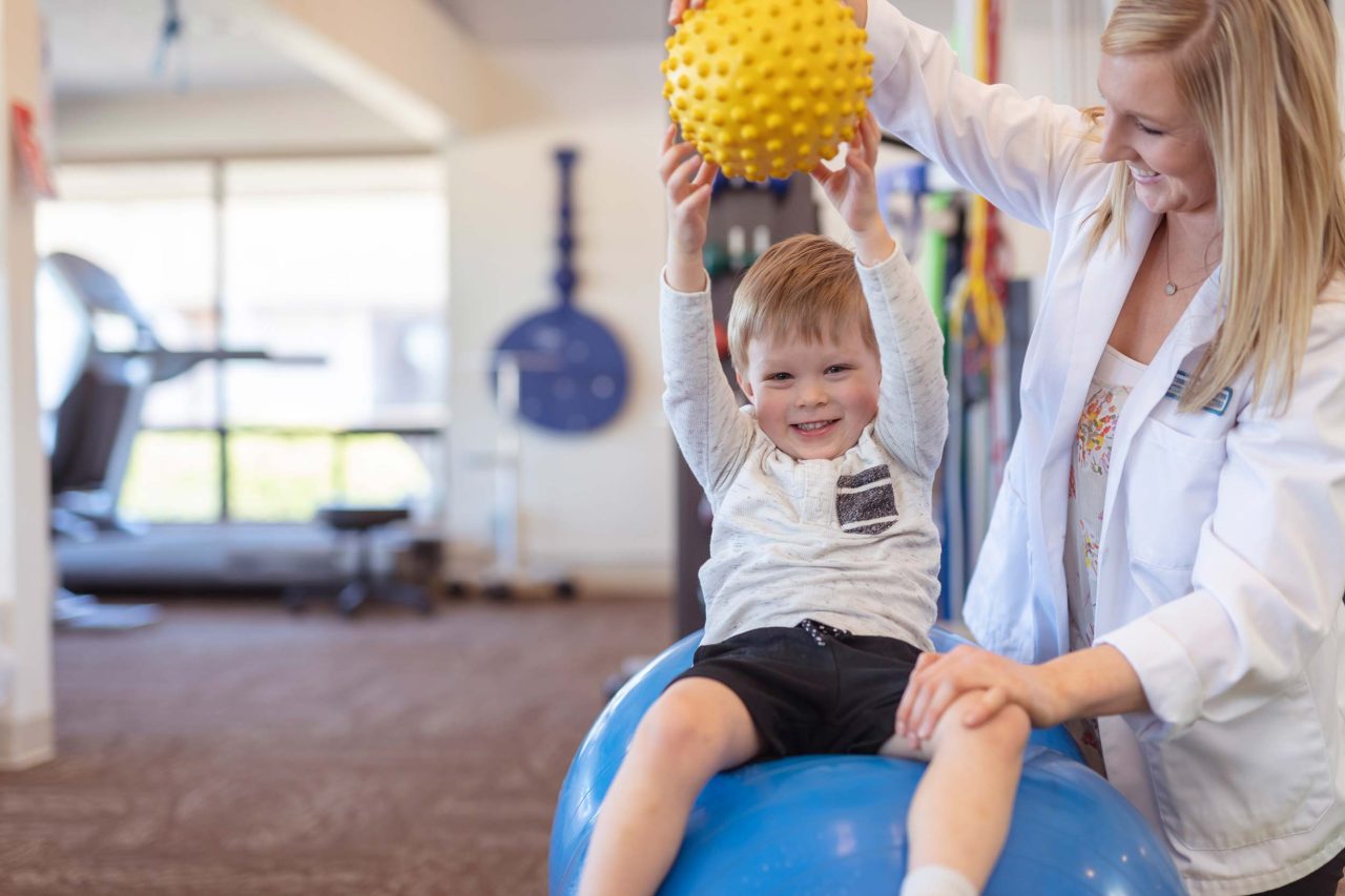 A smiling young boy holds a yellow ball above his head during a physical therapy session