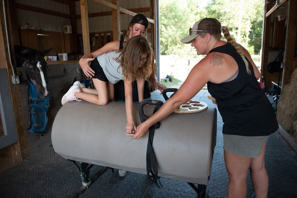 Two therapists assist a young child in a barn during a hippotherapy session