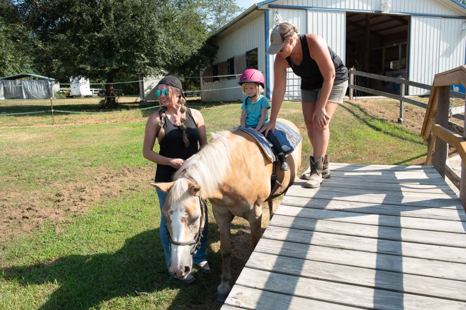 Two therapists assist a young girl on horseback during a hippotherapy session