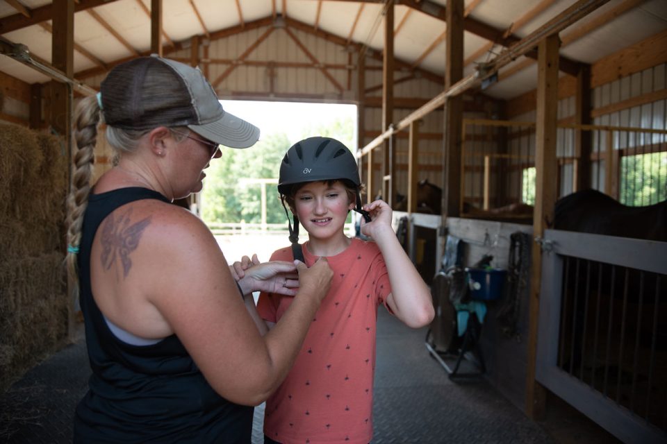 A therapist assists a young boy in putting on a helmet in a barn prior to a hippotherapy session